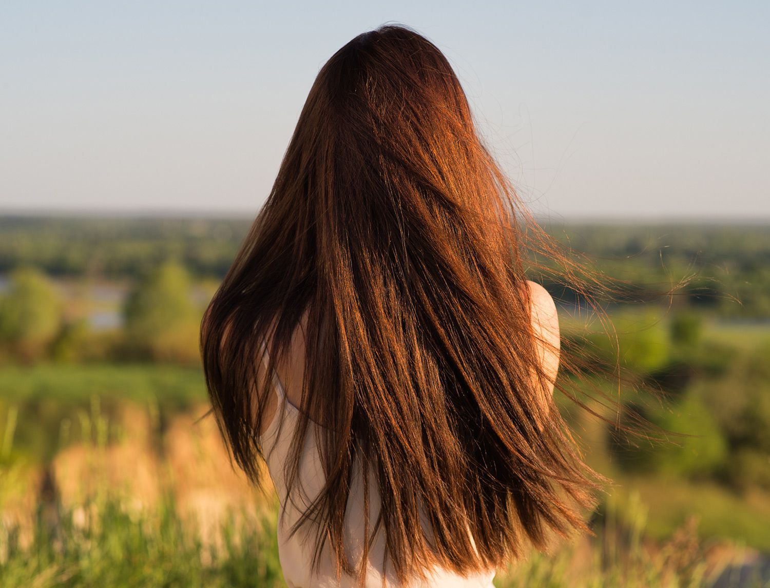 Donna vista di spalle con lunghi capelli castani mossi dal vento, su uno sfondo di campagna al tramonto