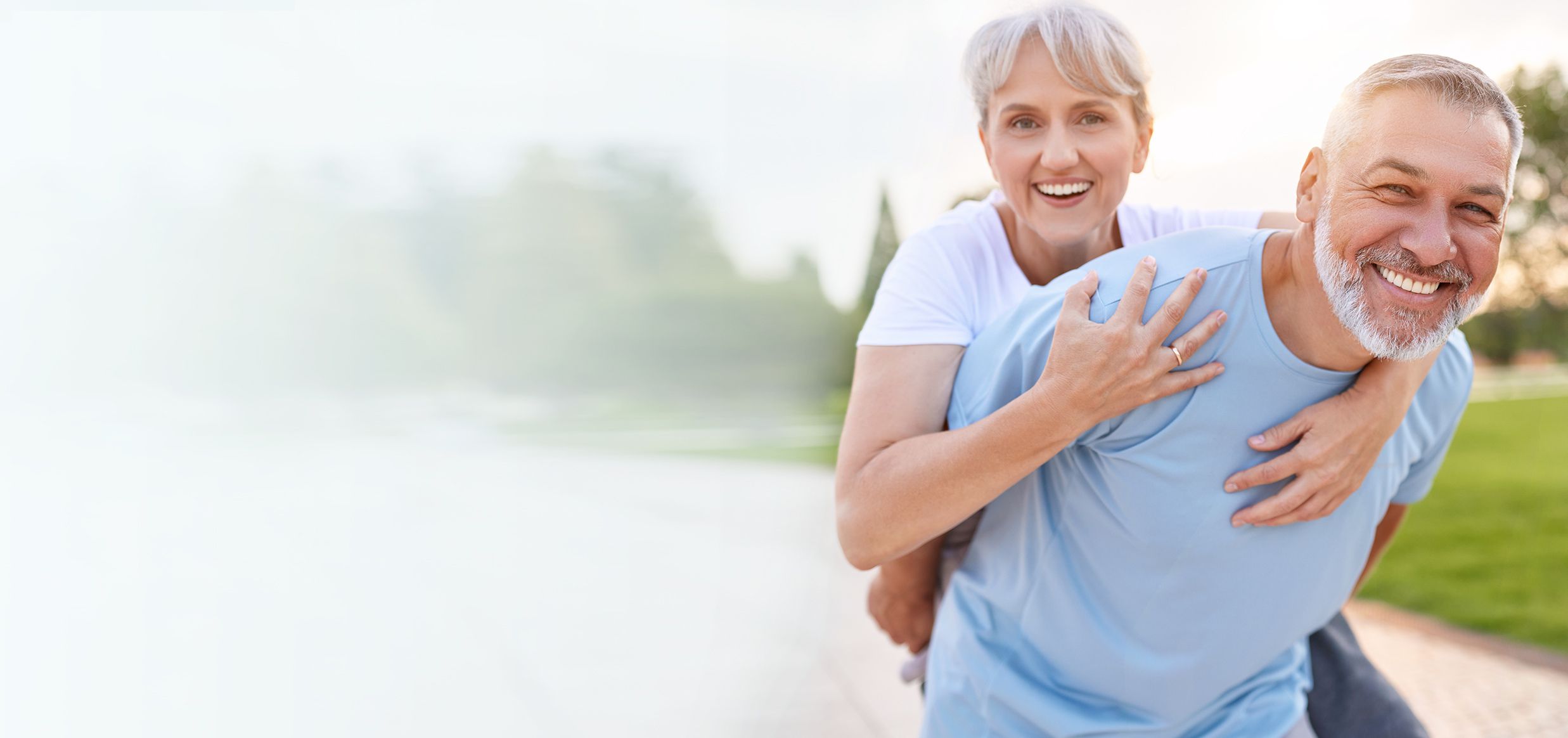 Coppia di anziani sorridenti che corrono abbracciati lungo un lago, lei sulle spalle di lui, in una giornata di sole