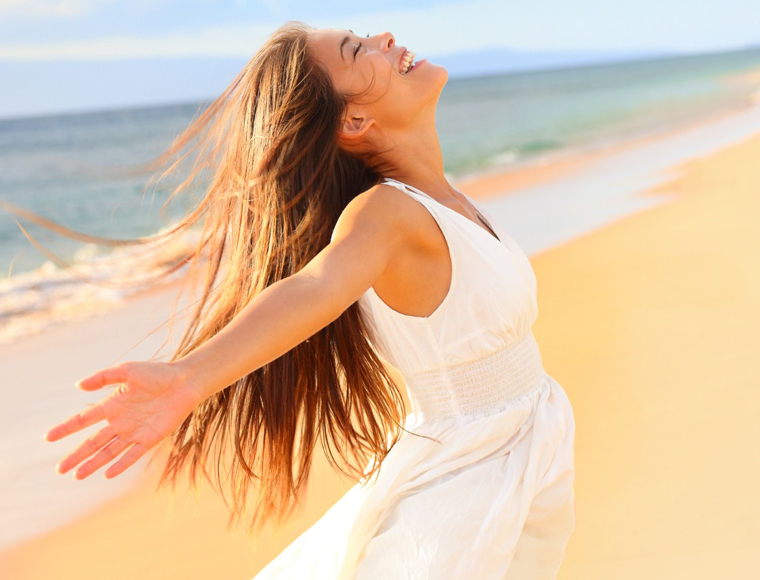 Donna con un abito bianco su una spiaggia, capelli al vento, braccia aperte e volto rivolto verso il cielo, sorridente