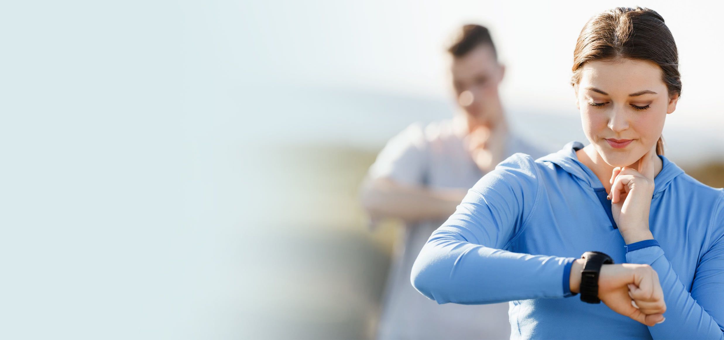 Donna in tenuta sportiva che controlla lo smartwatch al polso, con un uomo sfocato sullo sfondo