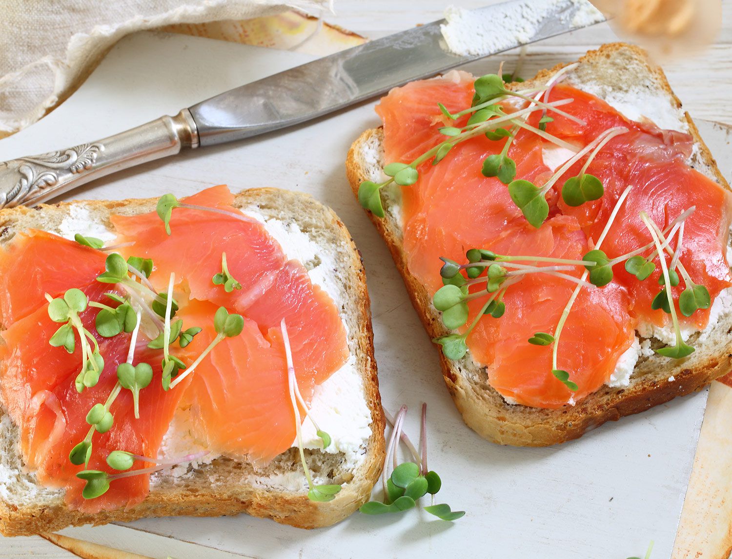 Due fette di pane tostato con crema di formaggio, salmone affumicato e germogli, su un tagliere bianco