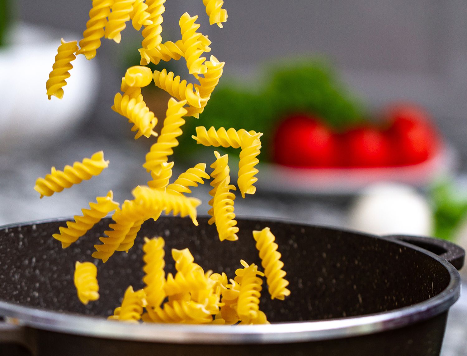 Fusilli crudi che cadono in una pentola scura, con pomodorini e uno spicchio d’aglio sfocati sullo sfondo