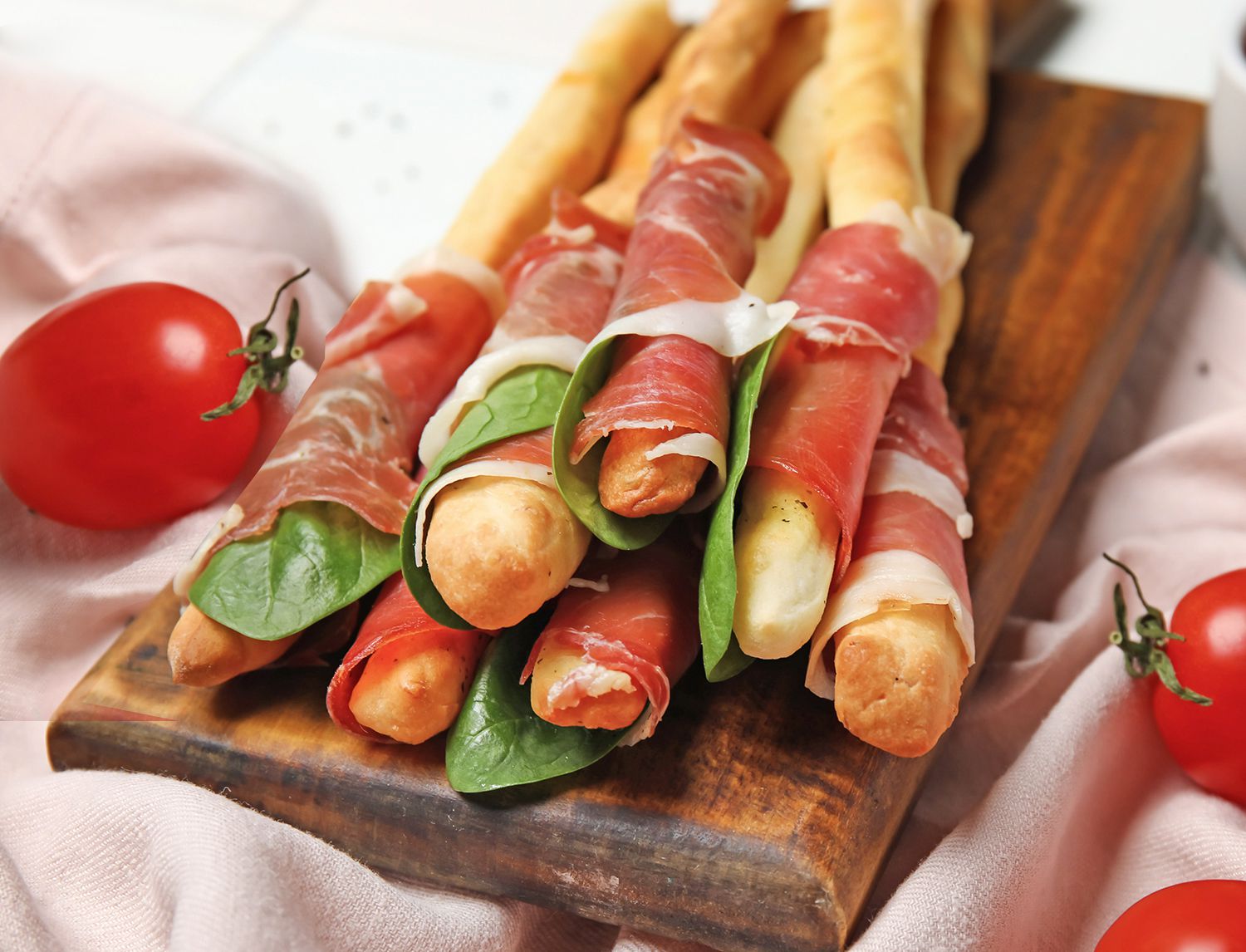 Grissini avvolti nel prosciutto crudo e in foglie di spinacino, disposti su un tagliere di legno con pomodorini