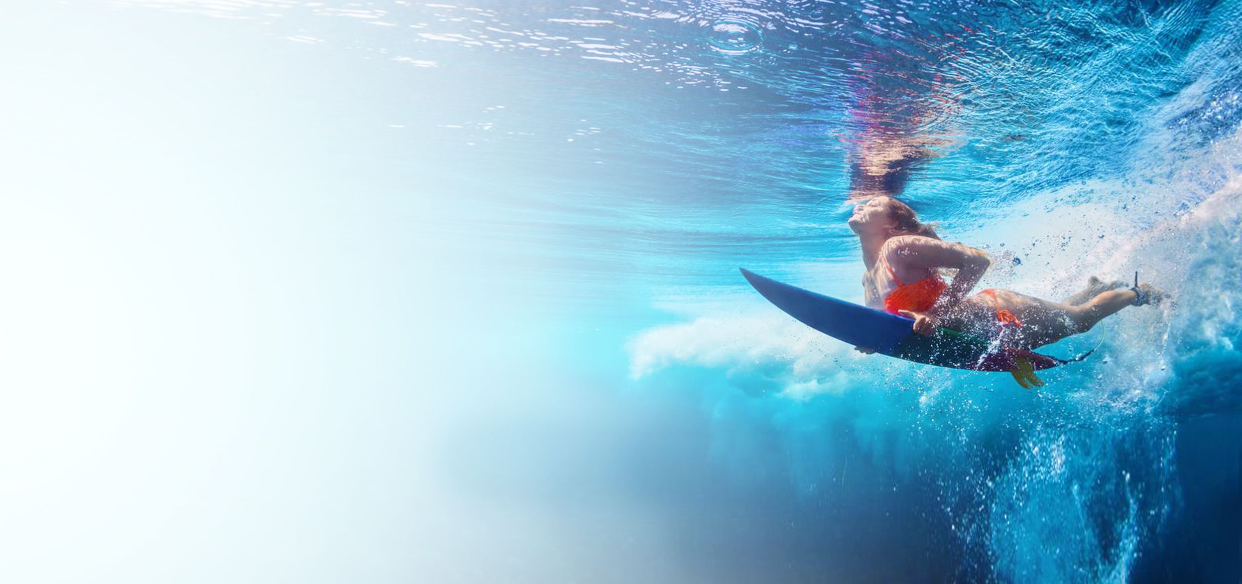 Persona che riemerge subacquea sotto un'onda con la tavola da surf, vista dal fondo verso la superficie illuminata