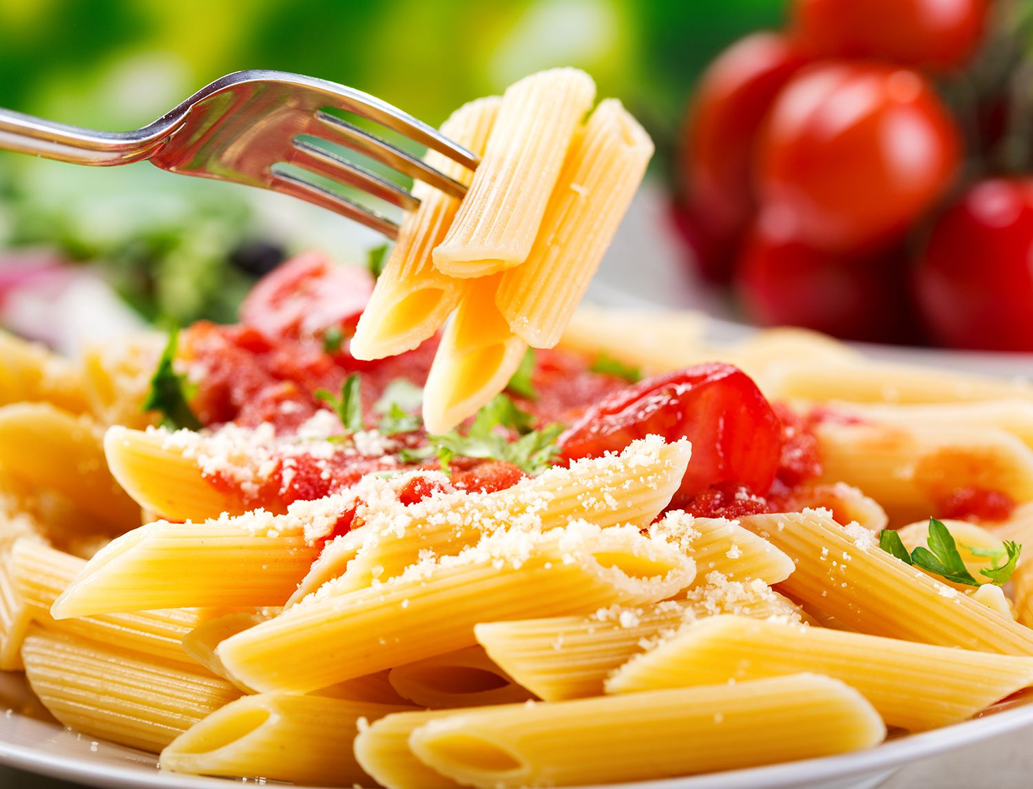 Forchetta che solleva penne al pomodoro condite con formaggio grattugiato e prezzemolo, con pomodorini sullo sfondo