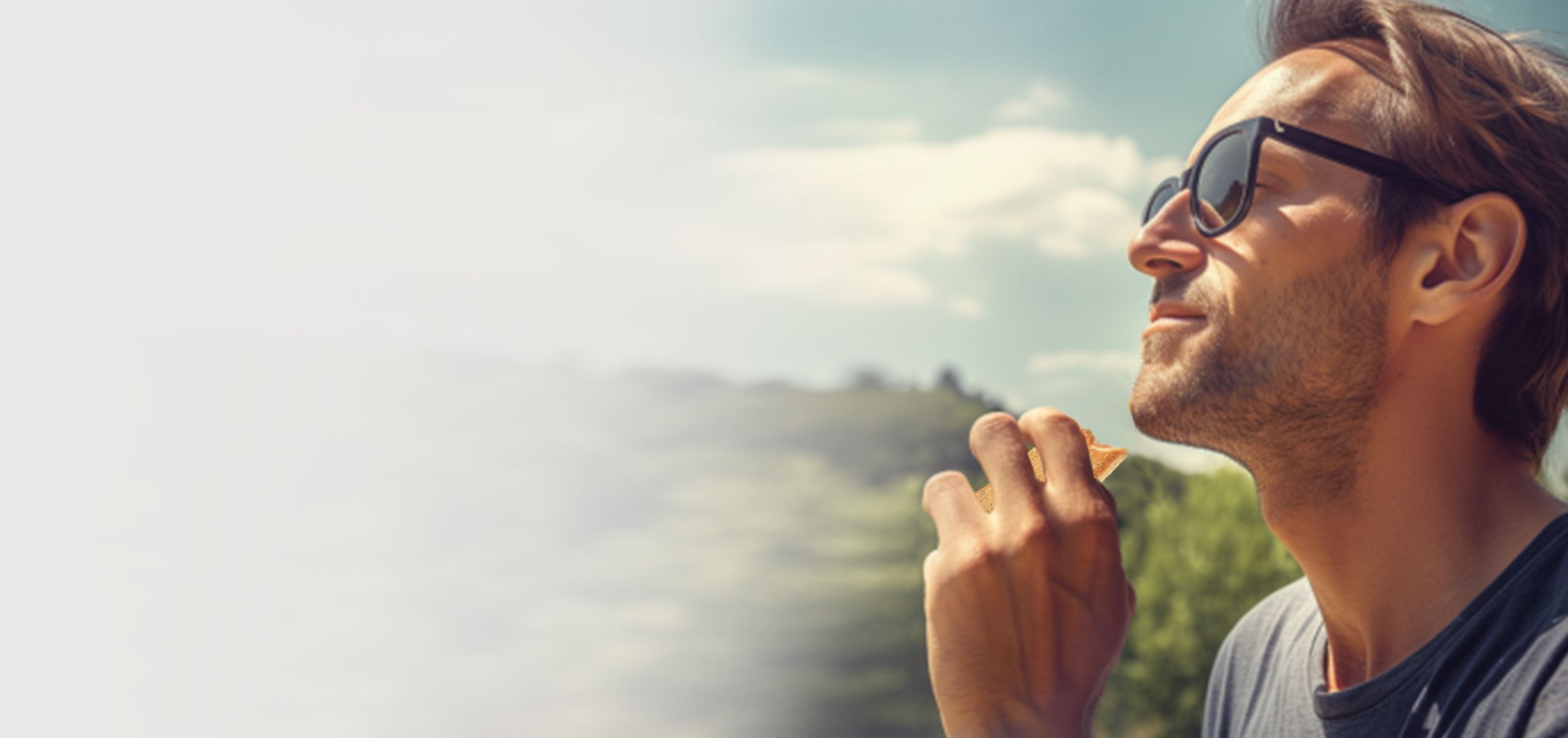 Uomo con occhiali da sole che mangia uno spuntino all’aperto, volto rivolto verso il sole, sfondo di colline verdi