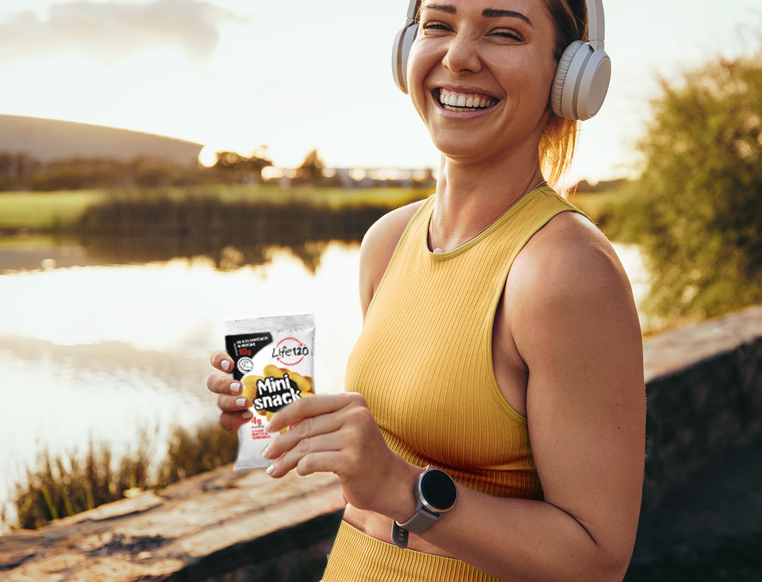 Donna con cuffie e abbigliamento sportivo che sorride tenendo in mano una confezione di Mini Snack, all’aperto vicino a un lago