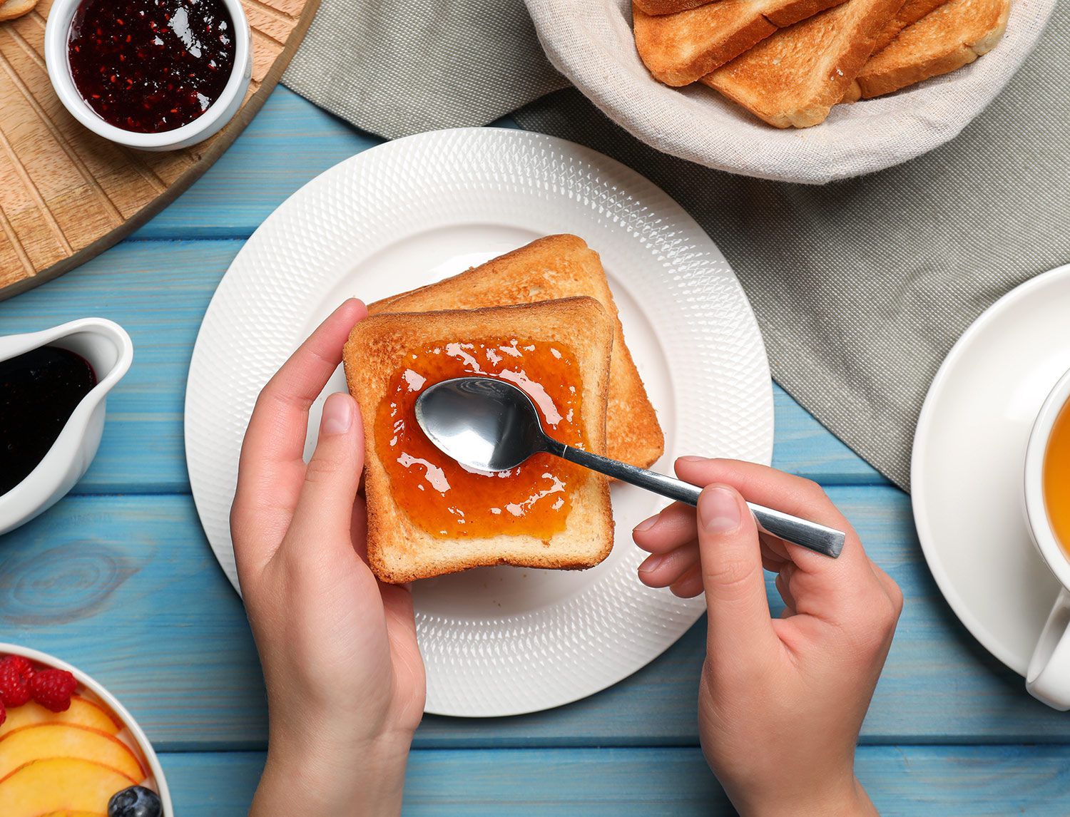 Mani che spalmano marmellata di albicocca su una fetta di pane tostato con un cucchiaino, tavola apparecchiata per colazione