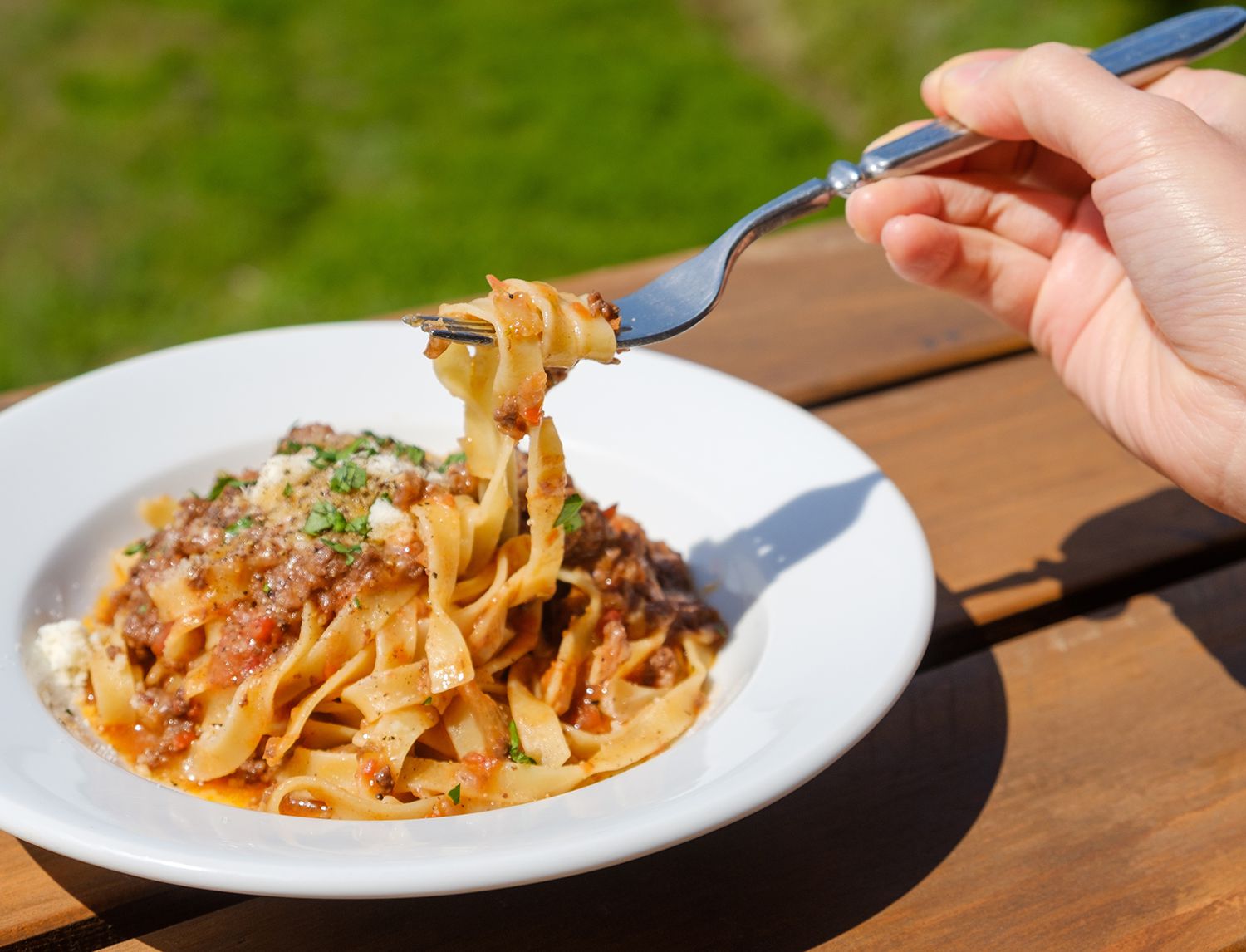 Forchetta che solleva fettuccine al ragù da un piatto bianco, all’aperto su un tavolo di legno