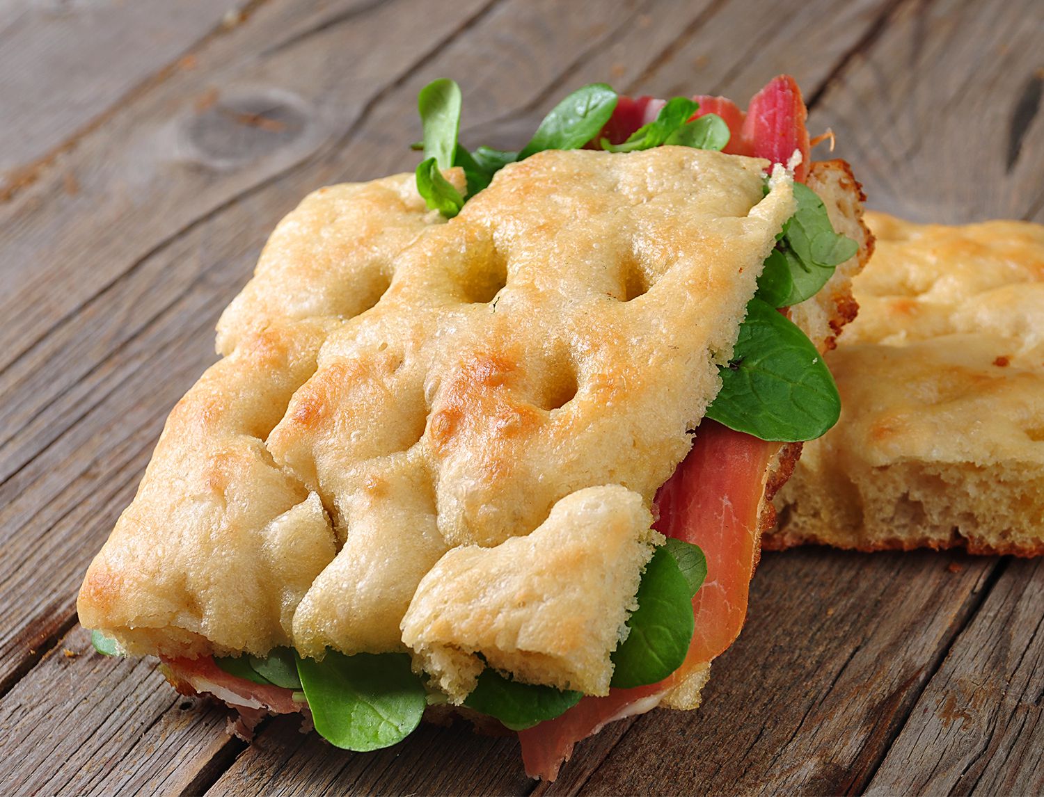 Primo piano di un panino di focaccia farcito con prosciutto crudo e valeriana, su un tavolo di legno