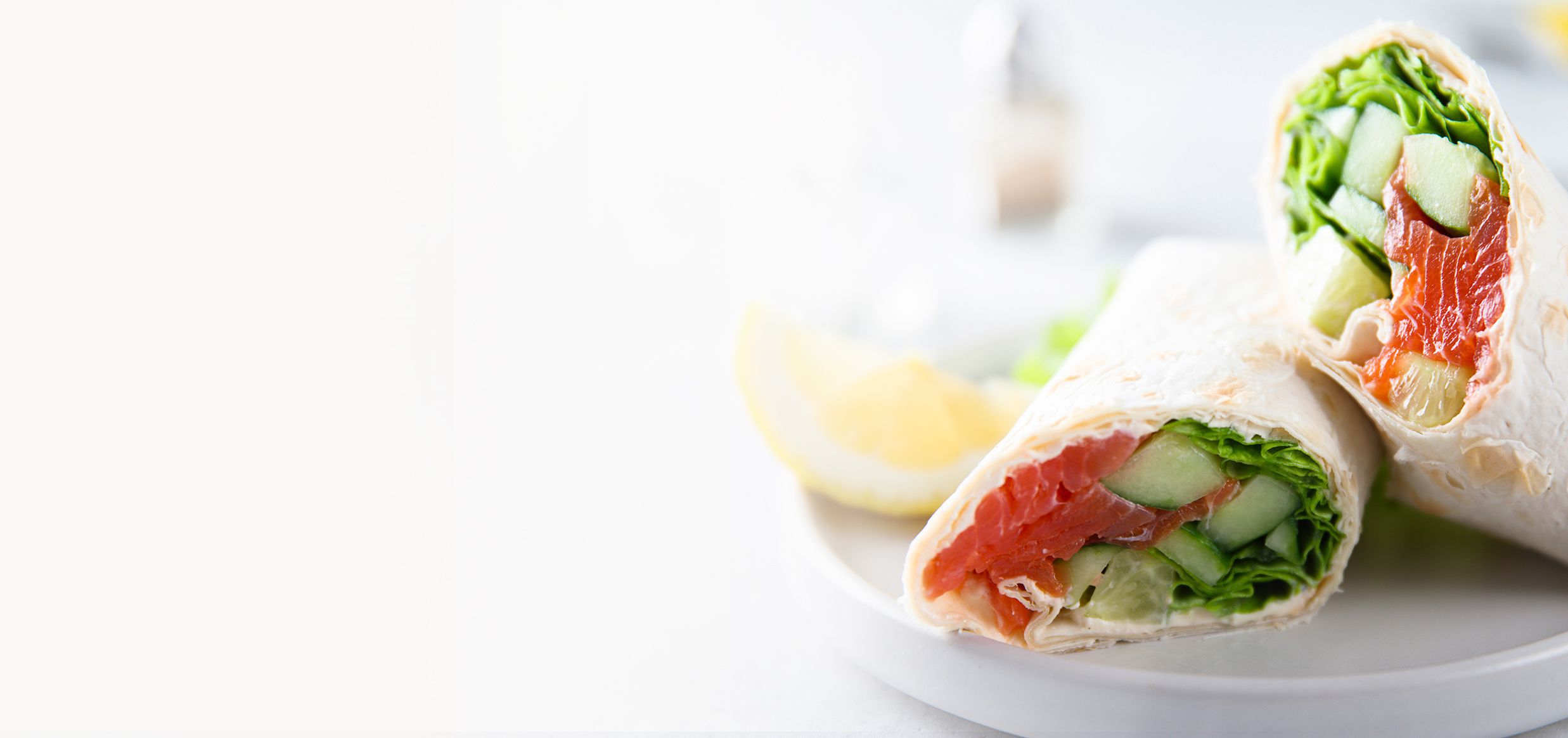 Due involtini di piadina farciti con salmone, cetriolo e insalata, tagliati a metà, su un piatto bianco
