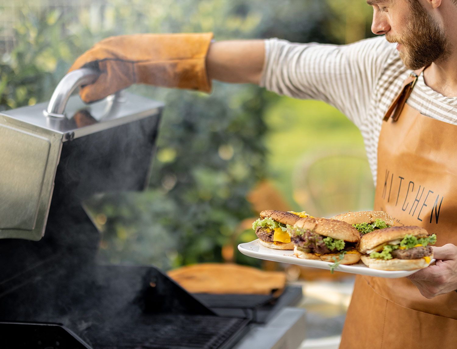 Uomo in grembiule da cucina che porta un piatto di hamburger farciti verso una griglia fumante, all’aperto