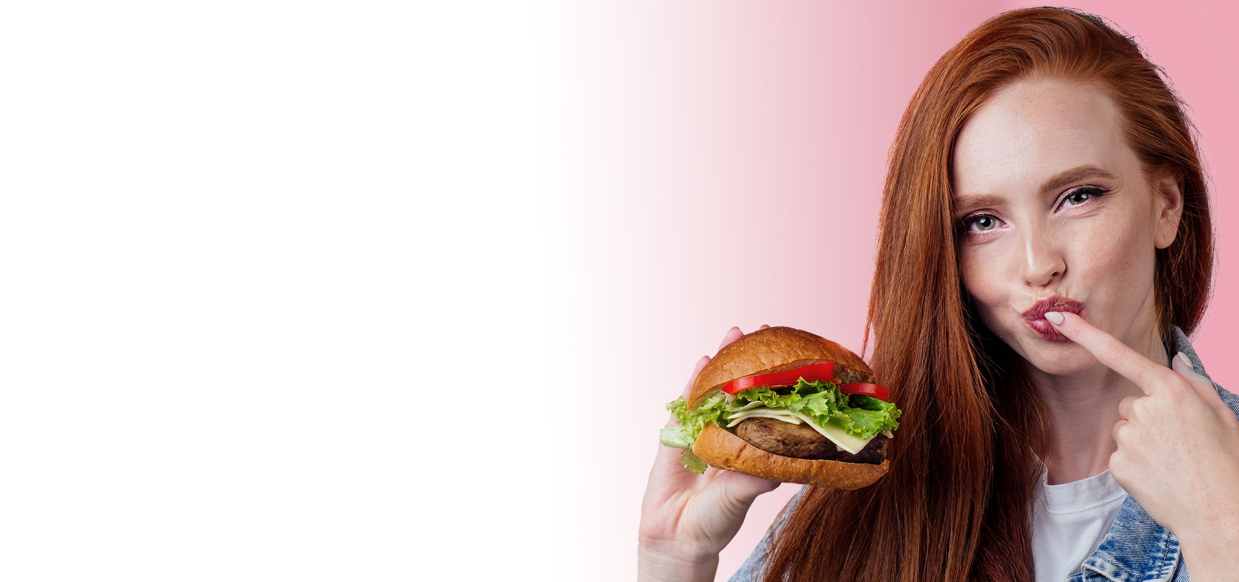 Giovane donna con i capelli rossi che morde un hamburger, con le dita vicino alla bocca, su sfondo rosa sfumato