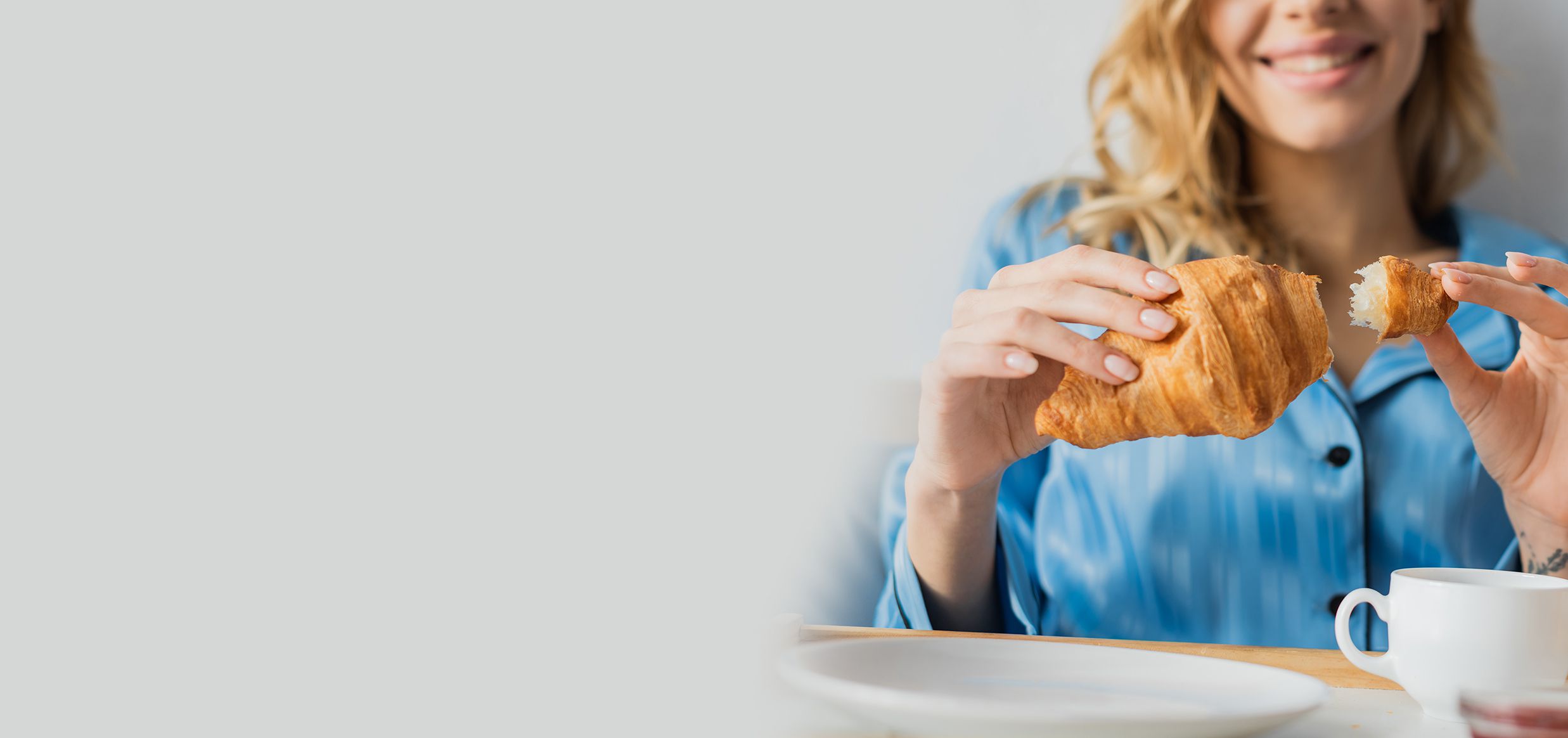 Donna in pigiama azzurro seduta a tavola che tiene in mano un cornetto morso, con una tazza bianca sul tavolo