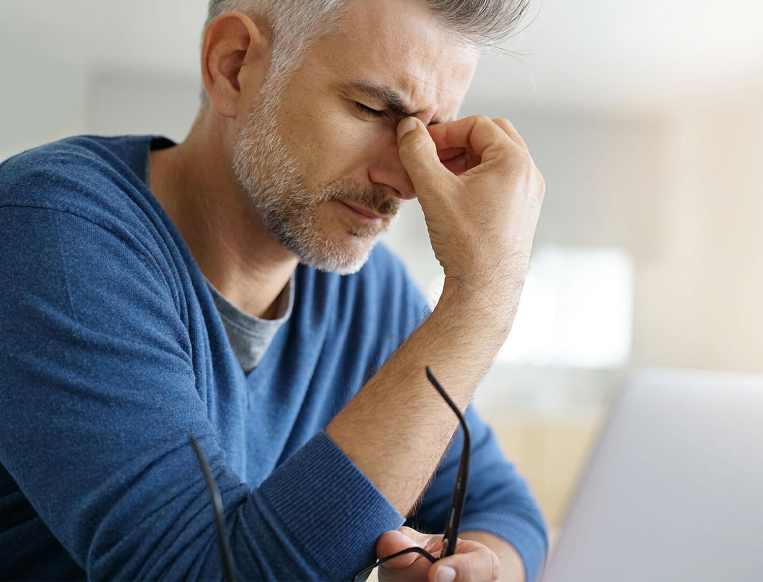 Uomo che si massaggia la fronte a occhi chiusi davanti a un laptop, con gli occhiali in mano — segno di stress o affaticamento