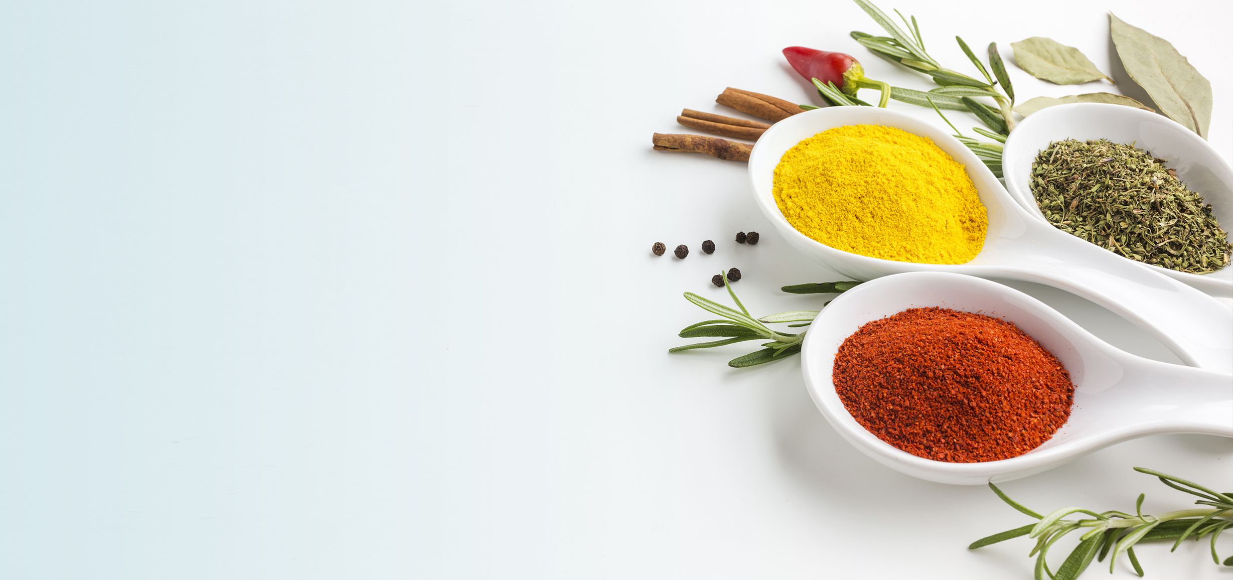 Tre cucchiai di ceramica con spezie in polvere (curcuma gialla, paprika rossa, origano essiccato), circondati da cannella, peperoncino, rametti di rosmarino e foglie di alloro