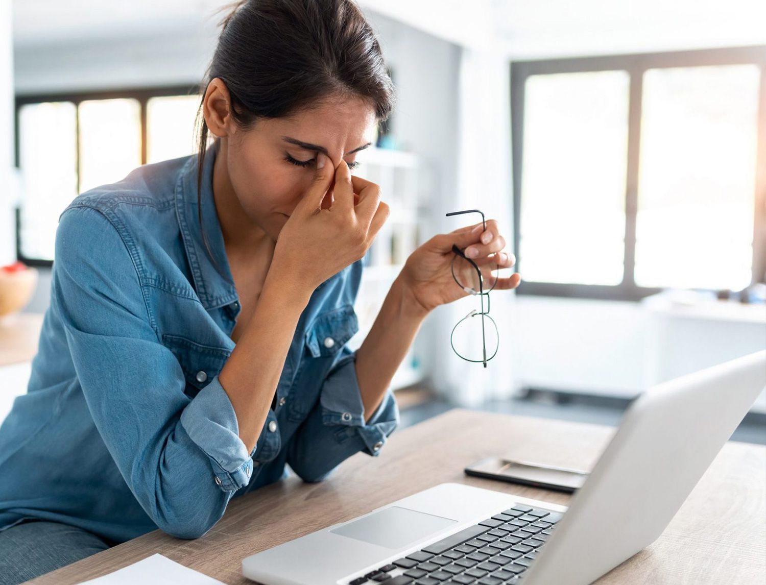 Donna alla scrivania con gli occhiali in mano che si massaggia il naso, davanti a un laptop aperto